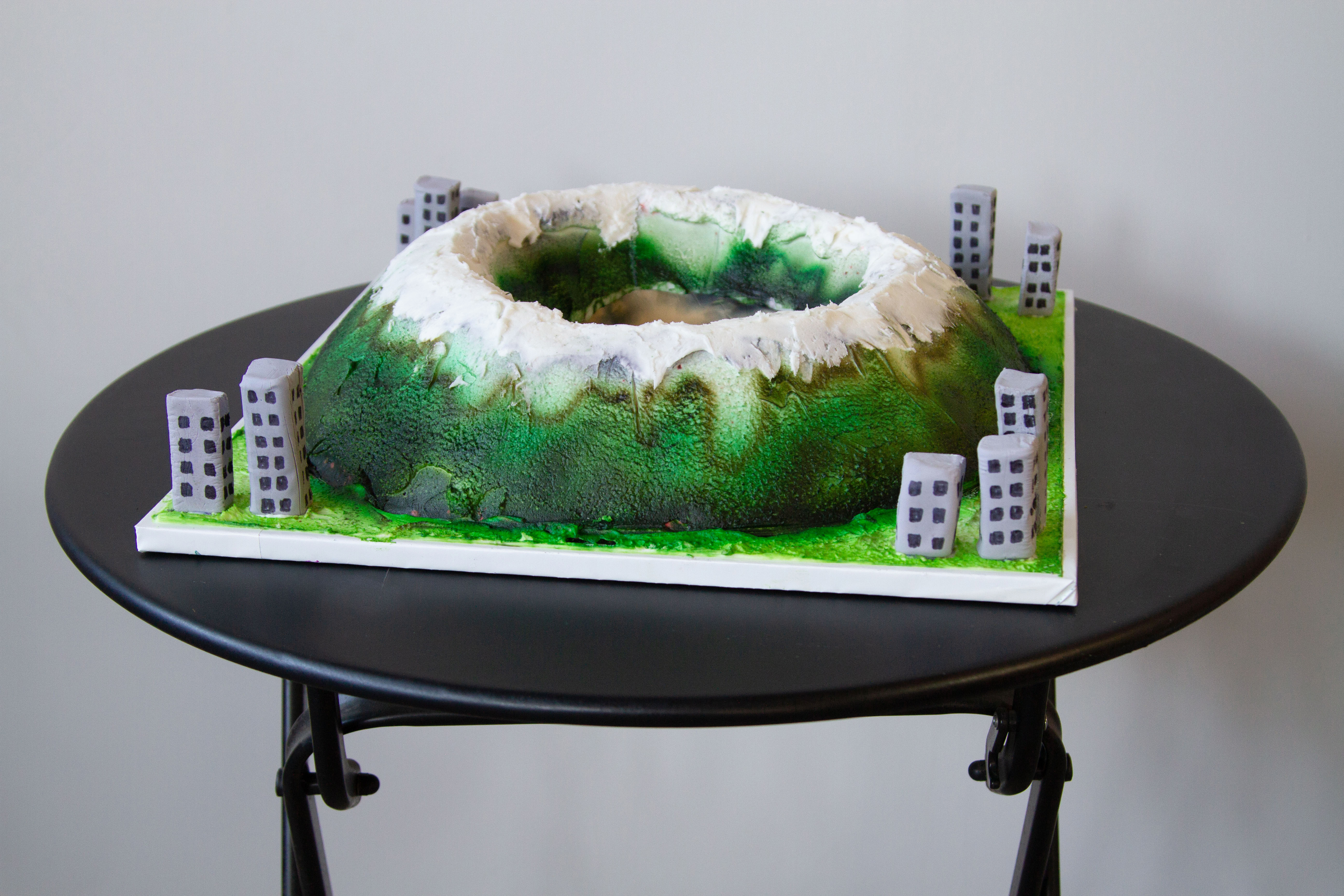 A cake shaped like a mountainous crater with buildings surrouding it on a black table